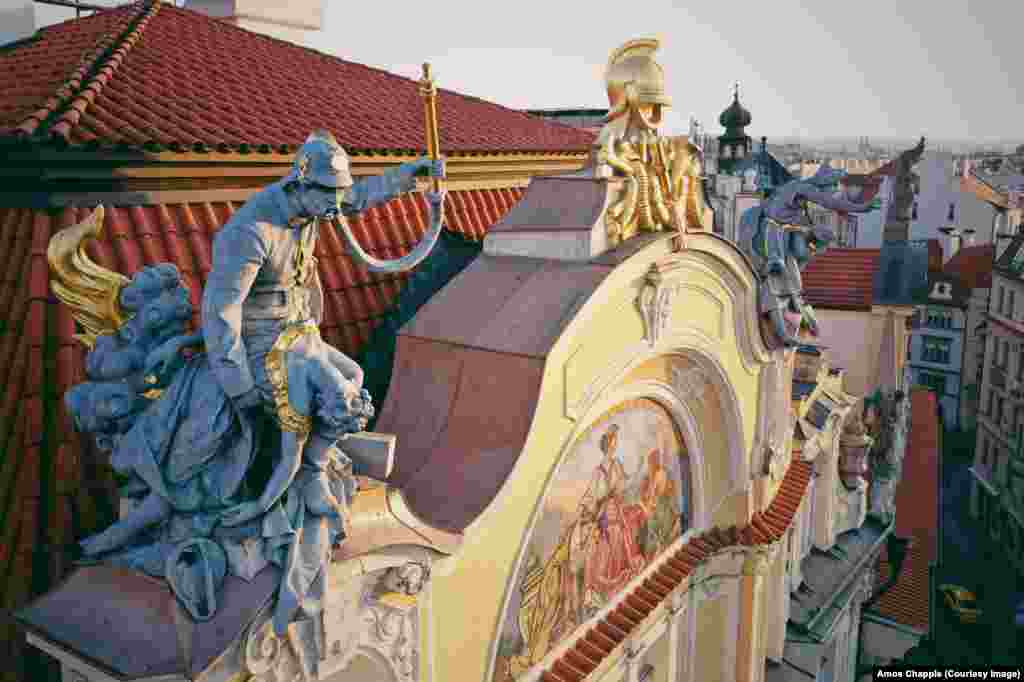 Пожежник рятує жінку від золотистого полум’я на даху будівлі колишньої Празької міської страхової компанії на Староміській площі в Празі. Ця драматична композиція була однією з останніх робіт чеського скульптора Богуслава Шнірха перед його смертю в 1901 році