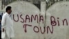 Pakistanly adam “Osama Bin Şäherçesi” diýlip, üstüne ýazgy ýazylan diwaryň deňinden geçip barýar, Abbottabat, 2011-nji ýylyň 6-njy maýy.